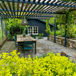 Ein gemütlicher Essbereich im Freien mit einem langen Holztisch und grünen Stühlen befindet sich unter einer Pergola neben einem schwarzen Gartenhaus. Im Vordergrund blühen gelbe Blumen, umgeben von Grün und Steinpflaster.
