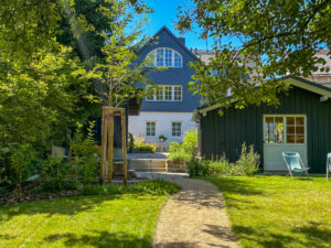 Ein sonnenbeschienener Garten mit einem gewundenen Weg führt zu einem charmanten blau-weißen Ferienhaus. Grüne Bäume und Sträucher rahmen die Szene ein, eine Terrasse mit Gartenmöbeln ist in der Nähe. Auf der rechten Seite vervollständigen ein grüner Schuppen und ein Liegestuhl diesen Rückzugsort in der Oberlausitz.
