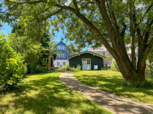Ein sonniger Garten mit einem geschwungenen Kiesweg, einem großen Baum und grünem Gras führt zu einem blauen Haus und einem kleinen schwarzen Gartenhaus mit zwei Stühlen im Freien. Diese friedliche Szenerie ist perfekt für einen entspannten Urlaub im Denkmal, umgeben von üppigem Grün.