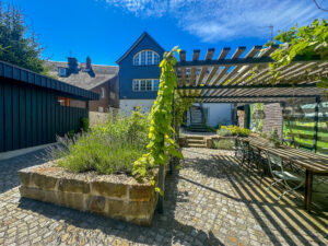 Ein sonniger Innenhof eines Ferienhauses mit Garten verfügt über eine mit Weinreben bewachsene Pergola, einen Esstisch im Freien mit Stühlen, mit Steinen eingefasste Pflanzbeete, Kopfsteinpflasterboden und ein Haus mit Schieferbehang und großen Fenstern im Hintergrund - das Marktquartier 5 in Neusalza-Spremberg