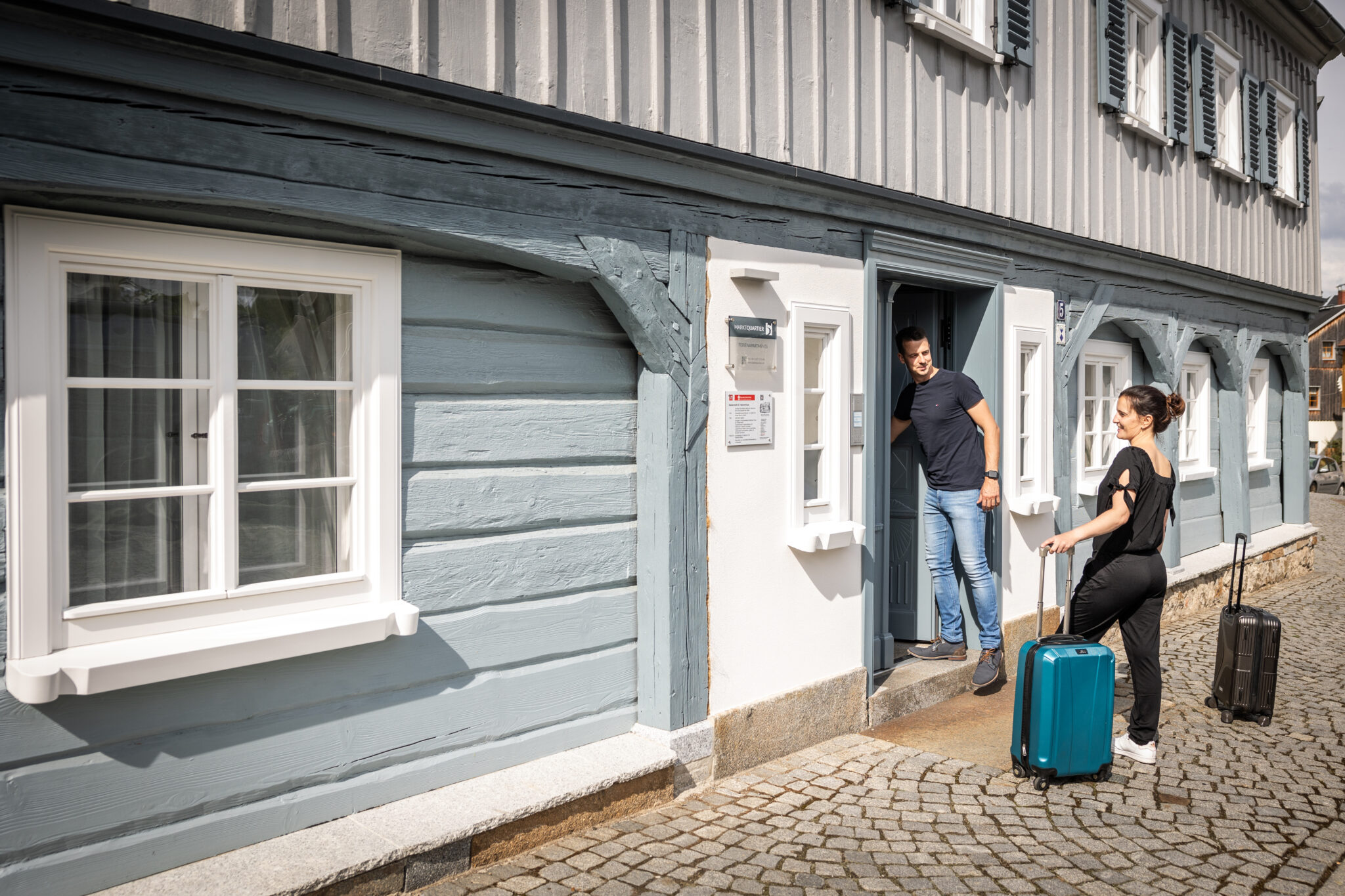 Ein Mann und eine Frau mit Rollkoffern stehen vor dem Eingang eines malerischen Holzhauses mit blau-weißen Wänden und Fensterläden. Die Frau lächelt, als der Mann die Tür aufhält – bereit für den Urlaub im Denkmal.