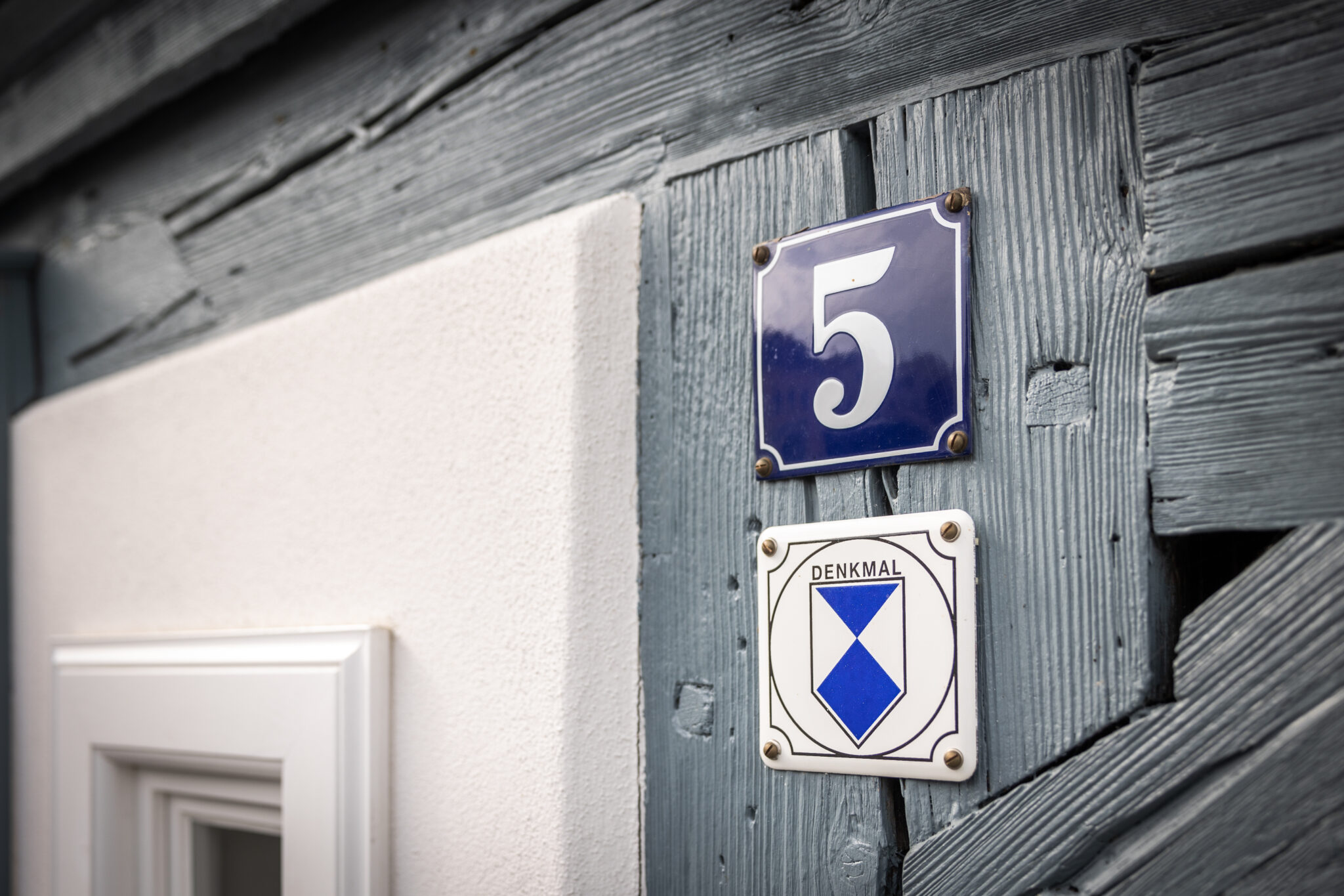 Blau-weiße Plakette mit der Hausnummer 5 über einem DENKMAL-Schild auf einer strukturierten Holzwand neben einem weißen Fensterrahmen, die auf den Charme von „Urlaub im Denkmal“ hinweist.