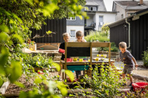 Drei kleine Kinder spielen an einem sonnigen Tag mit Spielzeug und malen an einem Holztisch im üppigen, grünen Garten eines Ferienhauses mit Garten, umgeben von Pflanzen und mit Häusern im Hintergrund.