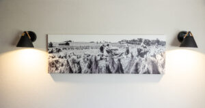 Ein Schwarz-Weiß-Foto von Menschen, die auf einem Feld arbeiten, hängt an einer weißen Wand zwischen zwei schwarzen, kegelförmigen Wandlampen, deren Licht auf das Kunstwerk scheint.