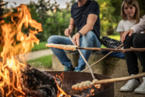 Zwei Personen sitzen am Lagerfeuer und rösten Teig auf Stöcken über den Flammen. Die Szene spielt im Freien mit grünem Hintergrund, und der Fokus liegt auf dem Lagerfeuer und dem Rösten.