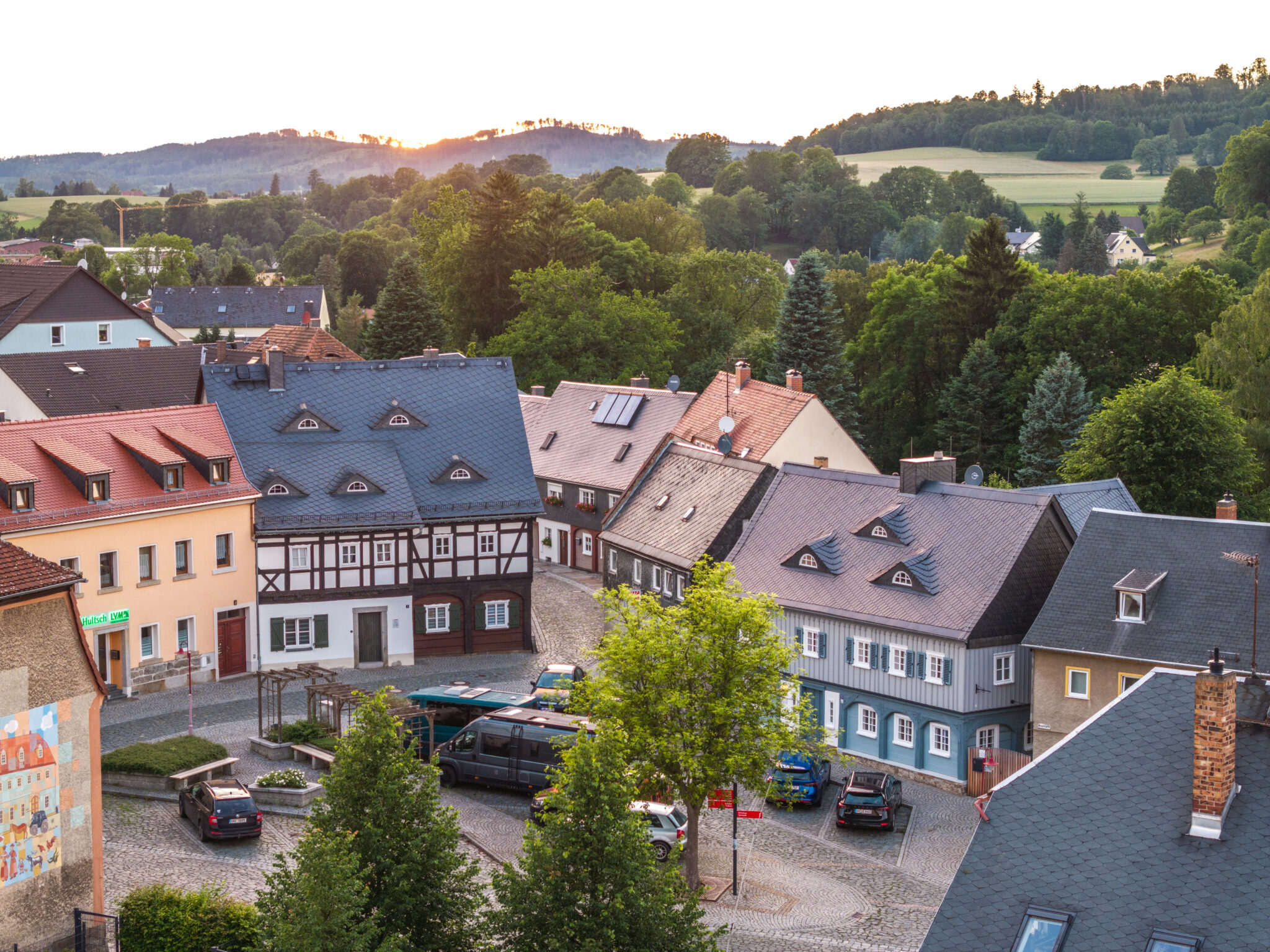 Eine malerische oberlausitzer Kleinstadt bei Sonnenuntergang mit bunten Fachwerkhäusern, Kopfsteinpflasterstraßen, geparkten Autos und üppigen grünen Bäumen in der Umgebung; im Hintergrund sind sanfte Hügel zu sehen.