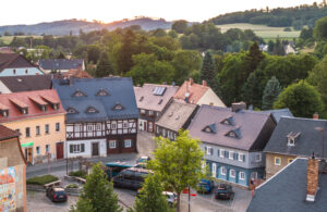 Eine malerische oberlausitzer Kleinstadt bei Sonnenuntergang mit bunten Fachwerkhäusern, Kopfsteinpflasterstraßen, geparkten Autos und üppigen grünen Bäumen in der Umgebung; im Hintergrund sind sanfte Hügel zu sehen.