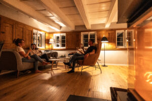 Vier Personen sitzen und unterhalten sich in einer gemütlichen, warm beleuchteten Ferienwohnung in der Oberlausitz mit Holzböden und -wänden in der Nähe eines Kamins. Die Atmosphäre ist entspannt und einladend, mit bequemen Stühlen und sanfter Beleuchtung.