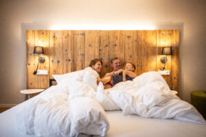 Eine glückliche dreiköpfige Familie - zwei Erwachsene und ein Kind - liegt gemeinsam unter weißen Decken auf einem Bett, lächelt und lacht in einem gemütlichen Schlafzimmer in ihrer Ferienwohnung in Neusalza-Spremberg im Marktquartier 5 und genießt die Wärme und den Komfort von Urlaub in der Oberlausitz.