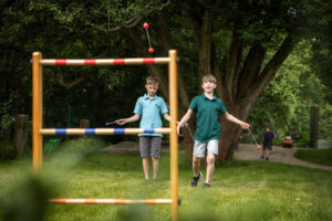 Zwei Jungen spielen Leiterwerfen auf einem Rasenplatz. Ein Junge wirft rote Bolas auf die Leiter, während der andere zusieht. Im Hintergrund sind Bäume und zwei weitere Kinder zu sehen.