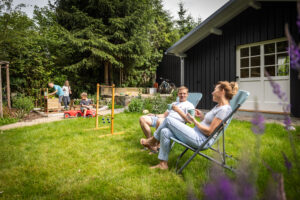 Ein Mann und eine Frau entspannen auf Gartenstühlen auf einer gepflegten Wiese, unterhalten sich und trinken Kaffee. Im Hintergrund spielen Kinder in der Nähe eines schwarzen Gartenhauses neben hölzernen Spielgeräten, umgeben von Bäumen und Grün. Der malerische Ort befindet sich direkt hinter dem Marktquartier 5, einem Ferienhaus mit Garten in Neusalza-Spremberg.