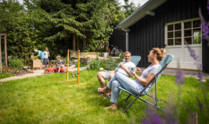 Ein Mann und eine Frau entspannen auf Gartenstühlen auf einer gepflegten Wiese, unterhalten sich und trinken Kaffee. Im Hintergrund spielen Kinder in der Nähe eines schwarzen Gartenhauses neben hölzernen Spielgeräten, umgeben von Bäumen und Grün. Der malerische Ort befindet sich direkt hinter dem Marktquartier 5, einem Ferienhaus mit Garten in Neusalza-Spremberg.