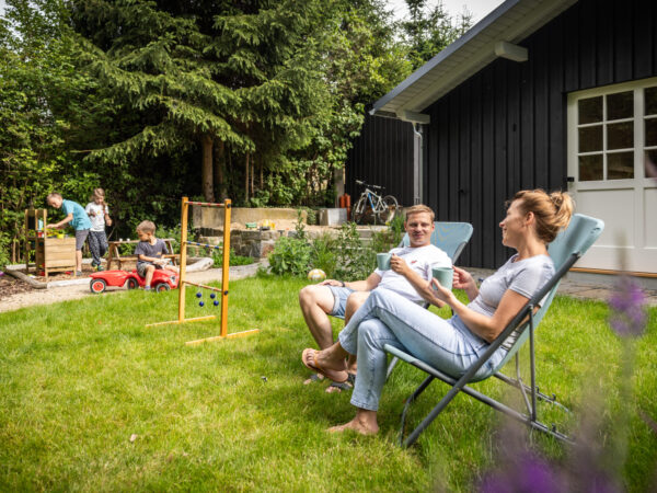 Ein Mann und eine Frau entspannen auf Gartenstühlen auf einer gepflegten Wiese, unterhalten sich und trinken Kaffee. Im Hintergrund spielen Kinder in der Nähe eines schwarzen Gartenhauses neben hölzernen Spielgeräten, umgeben von Bäumen und Grün. Der malerische Ort befindet sich direkt hinter dem Marktquartier 5, einem Ferienhaus mit Garten in Neusalza-Spremberg.