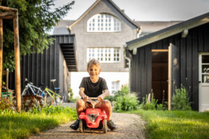 Ein lächelndes Kind fährt in einem kleinen roten Spielzeugauto auf einem Weg in einem sonnigen Hinterhof, mit modernen schwarzen Gebäuden und einem großen Haus im Hintergrund.