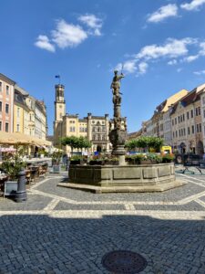Ein gepflasterter Stadtplatz mit Straßencafés, farbenfrohen historischen Gebäuden, einem zentralen Steinbrunnen mit einer Statue auf der Spitze und einem großen, kunstvollen Gebäude mit einem Uhrenturm im Hintergrund unter einem blauen Himmel. - Marktplatz Zittau mit Rathaus und Marsbrunnen. Ausflugsziel für den Urlaub in der Oberlausitz.
