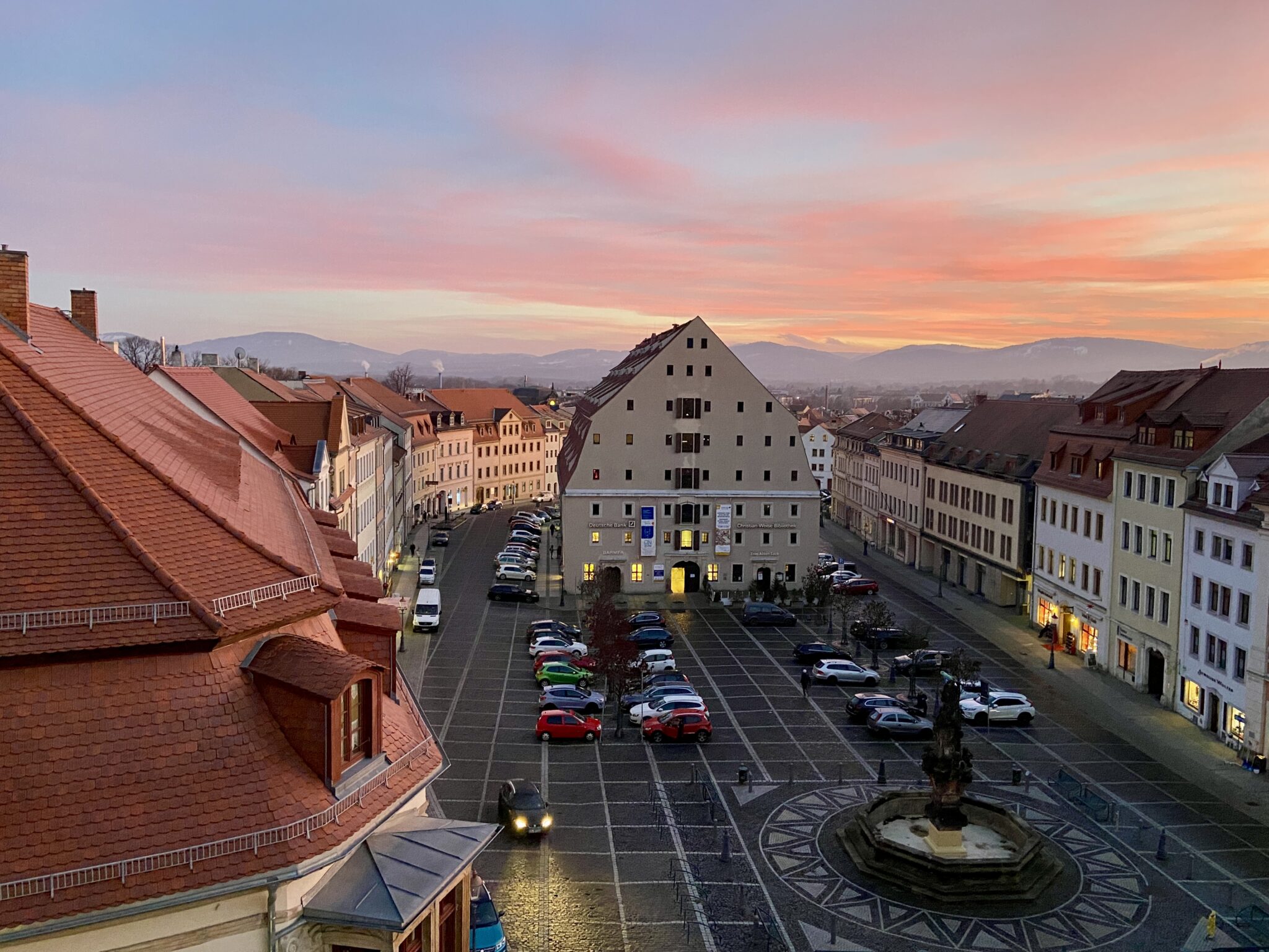 Ein europäischer Stadtplatz bei Sonnenuntergang mit geparkten Autos, einem Brunnen in der Mitte und historischen Gebäuden mit roten Dächern entlang der Straßen; der Himmel ist in zarte Rosa- und Orangetöne getaucht und in der Ferne sind Berge zu sehen (Zittau Neustadt mit Salzhaus).