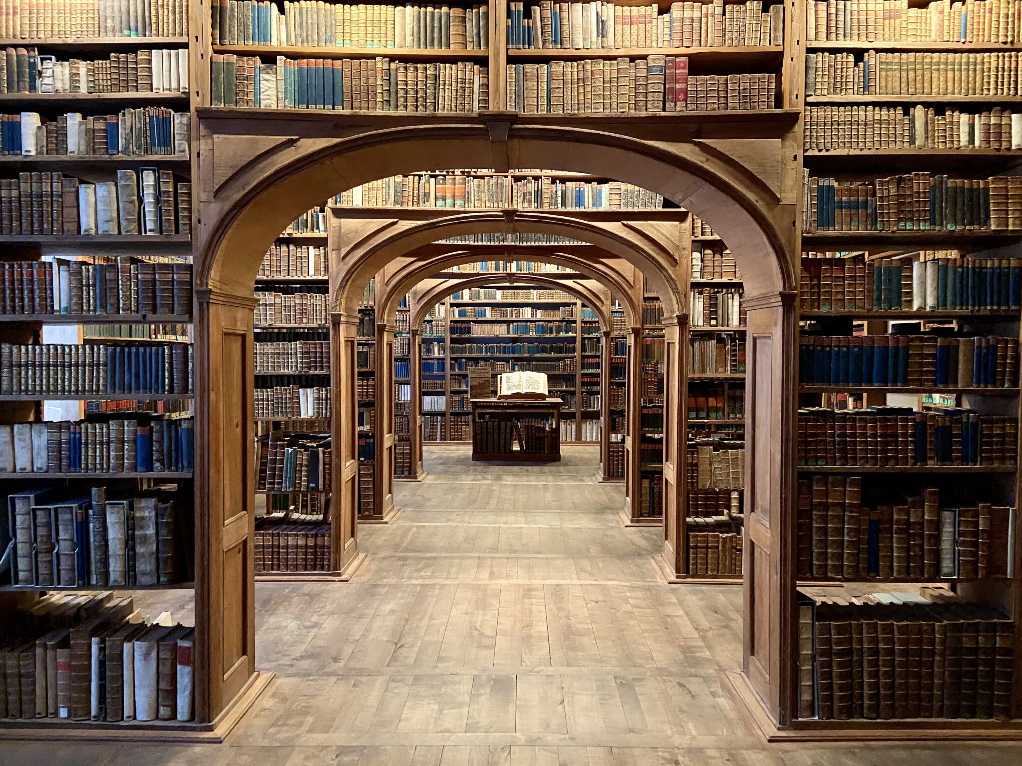 In einer großen, historischen Bibliothek reihen sich alte Bücher in hohen Holzregalen aneinander, die durch gewölbte Türen zu einem zentralen Tisch mit einem aufgeschlagenen Buch führen. Warmes Licht hebt das edle Holz und die alten Bände hervor. Wir sehen die Oberlausitzische Bibliothek der Wissenschaften in Görlitz. Lohnendes Ausflugsziel für den Urlaub in der Oberlausitz.
