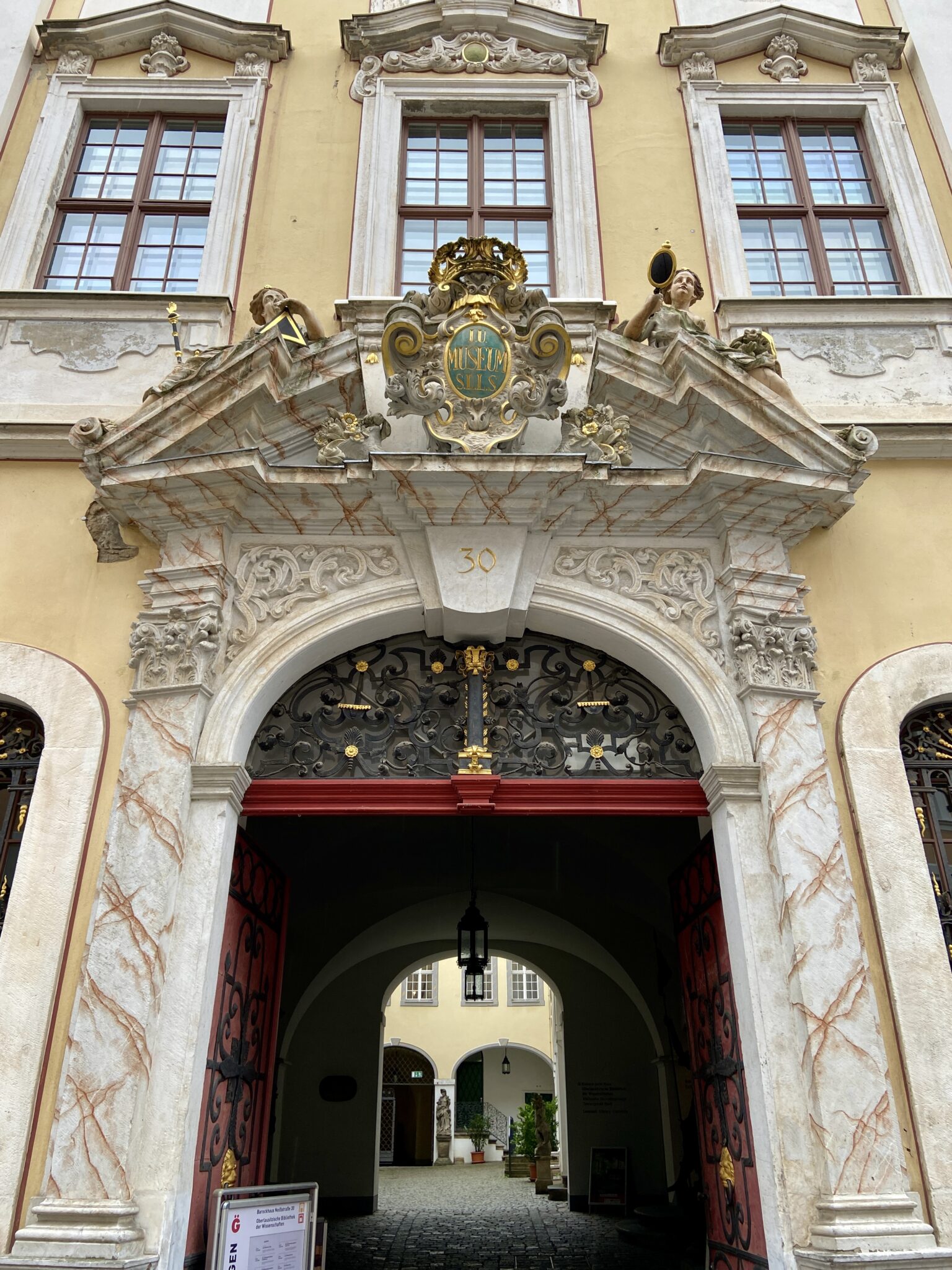 Der kunstvoll verzierte, gewölbte Eingang des Barockhauses in Görlitz mit Marmorsäulen, kunstvollen Schmiedearbeiten und goldenen Akzenten. Zwei Fenster im Obergeschoss und ein dekoratives Wappen mit Statuen und der Zahl 30 über dem Eingang.