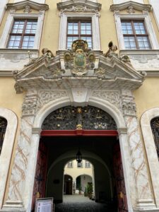 Der kunstvoll verzierte, gewölbte Eingang des Barockhauses in Görlitz mit Marmorsäulen, kunstvollen Schmiedearbeiten und goldenen Akzenten. Zwei Fenster im Obergeschoss und ein dekoratives Wappen mit Statuen und der Zahl 30 über dem Eingang.