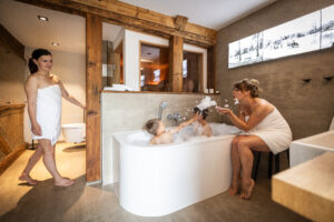Zwei Frauen und zwei Kinder befinden sich in einem modernen Badezimmer. Eine Frau steht mit einem Handtuch bekleidet da, während die andere neben einer Badewanne sitzt und Seifenblasen für die beiden Kinder macht, die baden und mit Seifenblasen spielen. Das Badezimmer befindet sich in der Ferienwohnung für große Familien im Marktquartier 5 in Neusalza-Spremberg.