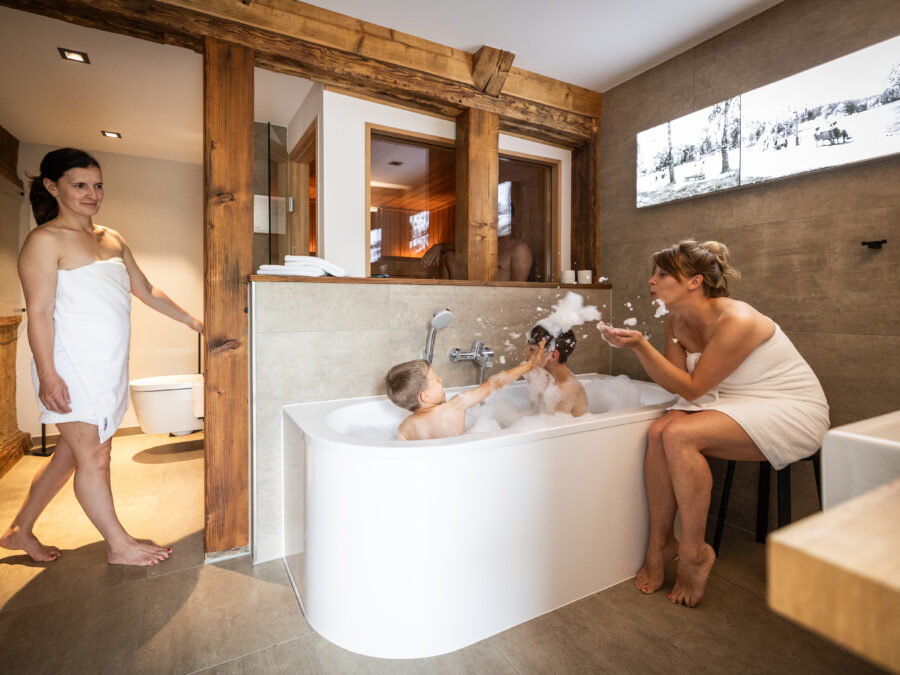 Zwei Frauen und zwei Kinder befinden sich in einem modernen Badezimmer. Eine Frau steht mit einem Handtuch bekleidet da, während die andere neben einer Badewanne sitzt und Seifenblasen für die beiden Kinder macht, die baden und mit Seifenblasen spielen. Das Badezimmer befindet sich in der Ferienwohnung für große Familien im Marktquartier 5 in Neusalza-Spremberg.