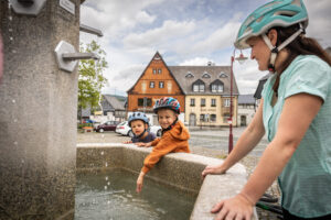 Eine Frau und zwei Kinder mit Fahrradhelmen stehen an einem Steinbrunnen auf einem Stadtplatz, dem Obermarkt von Neusalza-Spremberg. Die Kinder lächeln und berühren das Wasser. Im Hintergrund sind historische Gebäude unter einem bewölkten Himmel zu sehen.