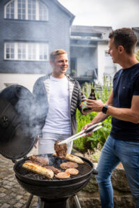 Zwei Männer stehen lächelnd und mit Bierflaschen in den Händen an einem Holzkohlegrill im Freien. Einer grillt Würstchen und Steaks mit einer Grillzange, Rauch steigt vom Grill auf. Im Hintergrund ist das Ferienhaus Marktquartier 5 zu sehen.