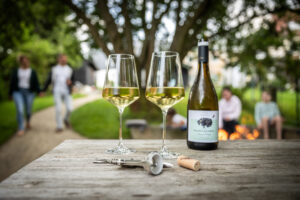 Zwei Gläser Weißwein und eine Weinflasche stehen auf einem Holztisch im Freien. Korken und Korkenzieher liegen daneben. Im Hintergrund genießen vier Personen die Gartenatmosphäre. Zwei gehen spazieren, zwei sitzen an einer Feuerstelle.