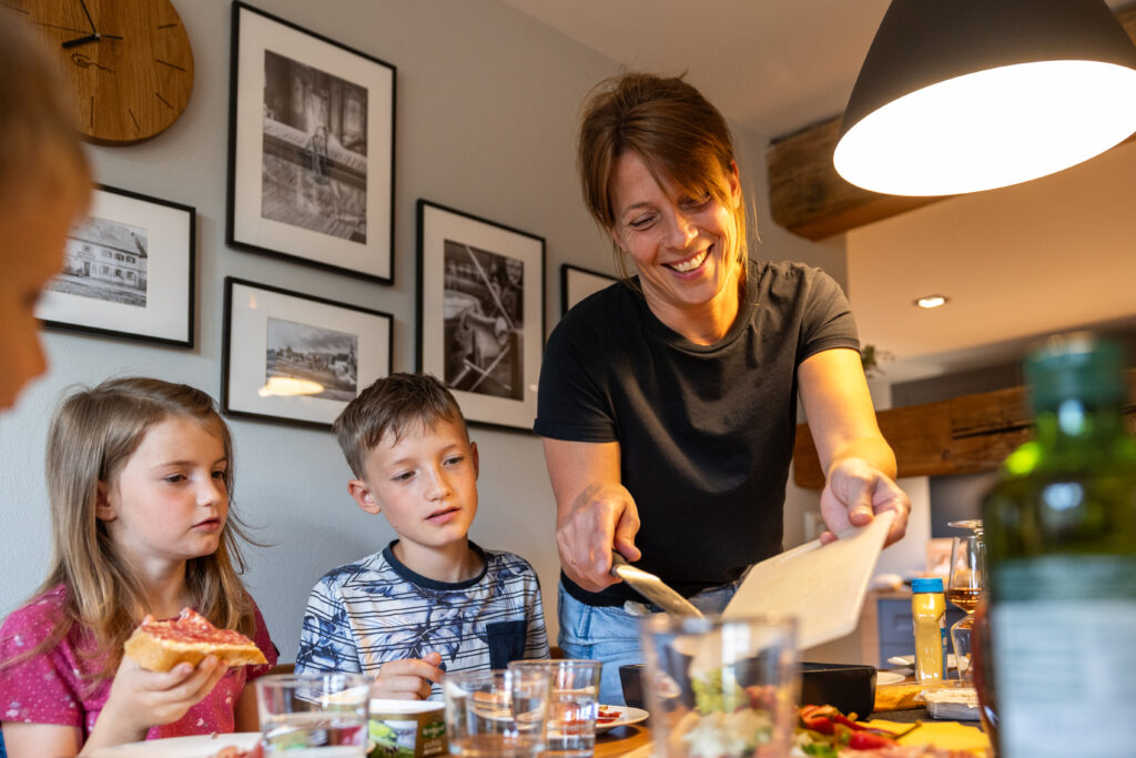 Eine Frau serviert zwei Kindern am Esstisch Essen. Die Kinder sitzen nebeneinander, eines isst eine Scheibe Brot. Die Stimmung ist fröhlich, die Atmosphäre ist gemütlich, modern, mit gerahmten Fotos an der Wand. Essbereich in einer Ferienwohnung für große Familien im Marktquartier 5 in Neusalza-Spremberg.