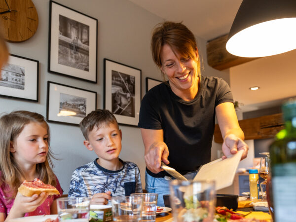 Eine Frau serviert zwei Kindern am Esstisch Essen. Die Kinder sitzen nebeneinander, eines isst eine Scheibe Brot. Die Stimmung ist fröhlich, die Atmosphäre ist gemütlich, modern, mit gerahmten Fotos an der Wand. Essbereich in einer Ferienwohnung für große Familien im Marktquartier 5 in Neusalza-Spremberg.