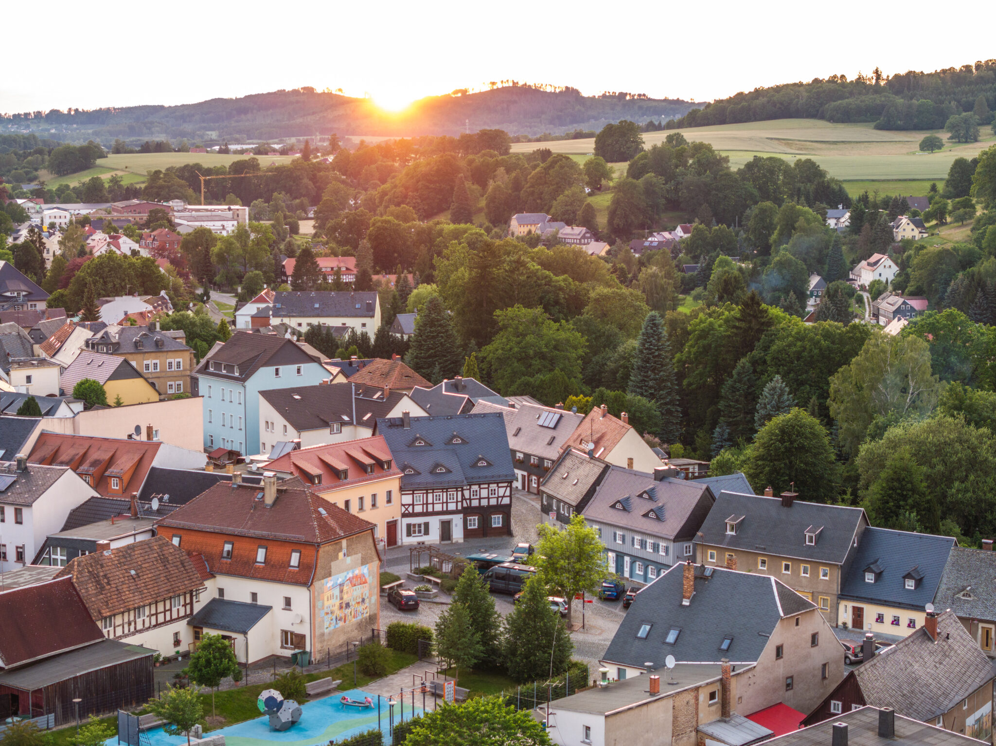 Ein malerischer Blick auf eine kleine Stadt mit bunten Häusern, Bäumen und sanften Hügeln bei Sonnenuntergang. Die Sonne versinkt hinter den Hügeln und wirft einen warmen Schein – perfekt für alle, die charmante Ferienwohnungen in Neusalza-Spremberg suchen.