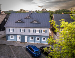 Ein hellblau-graues Holzhaus mit Dachgauben liegt an einer Kopfsteinpflasterstraße. Zwei Autos, ein blaues und ein schwarzes, parken davor. Grüne Bäume rahmen die Szene unter einem leicht bewölkten Himmel ein.