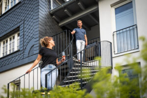 Eine Frau mit einem Glas in der Hand steht auf einer Wendeltreppe im Freien. Sie blickt nach oben und lächelt einem Mann zu, der über ihr am Geländer lehnt. Sie befinden sich vor einem modernen Gebäude mit großen Fenstern.