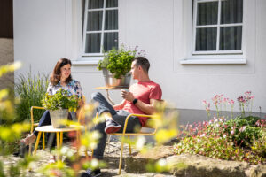 Eine Frau und ein Mann sitzen auf gelben Stühlen in einem Garten, unterhalten sich und trinken aus Tassen. Topfpflanzen stehen auf kleinen Tischen daneben, und in der Nähe blühen bunte Blumen. Im Hintergrund ist ein weißes Haus mit großen Fenstern zu sehen.