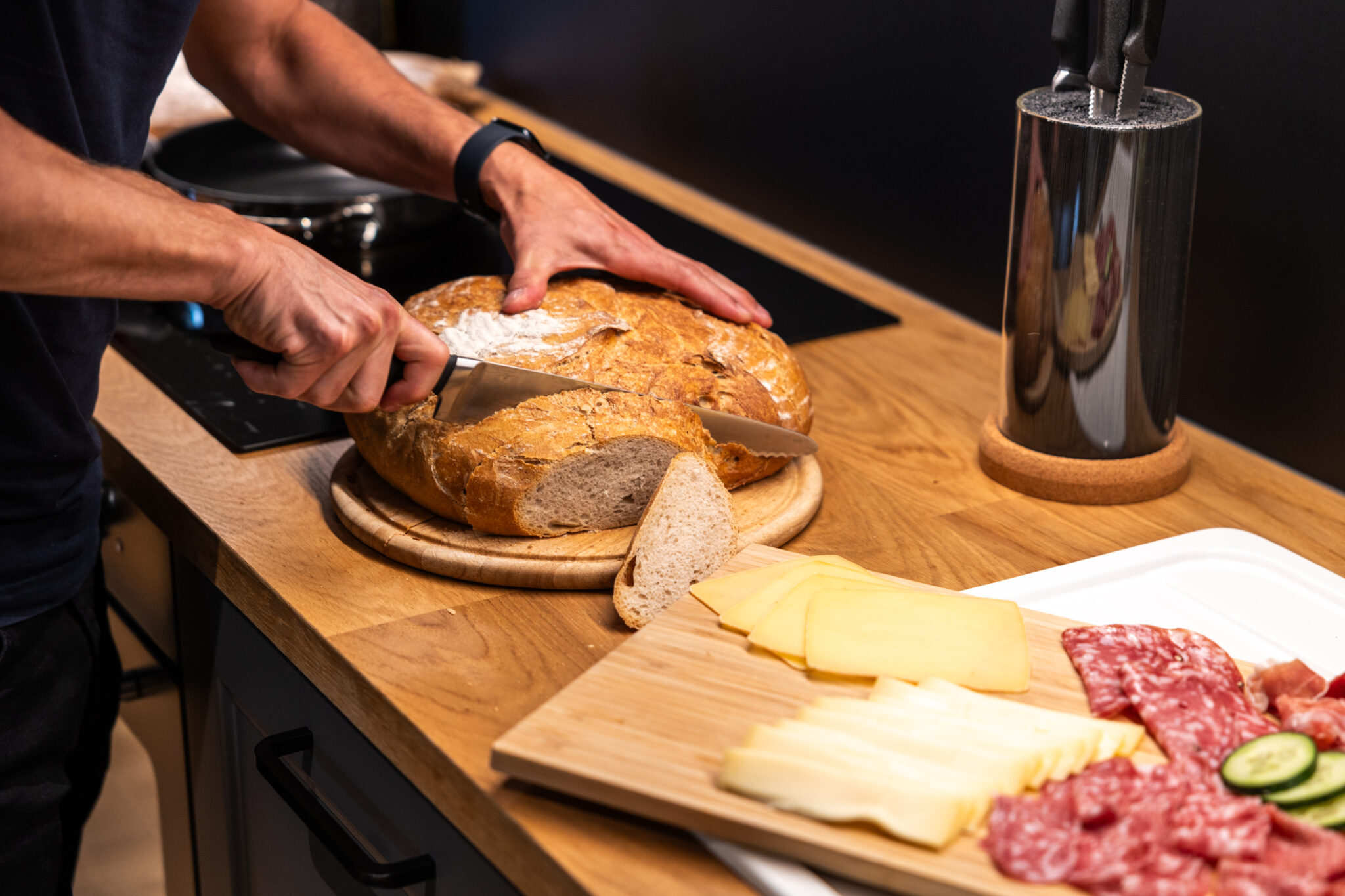 Eine Person schneidet in einer Küche einen runden Laib Brot auf einem hölzernen Schneidebrett. Daneben liegt ein Teller mit verschiedenen Käsesorten, Salami und Gurkenscheiben, und auf der Arbeitsplatte liegt ein Messerblock. Küche in einer Ferienwohnung für große Familien im Marktquartier 5 in Neusalza-Spremberg.