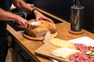 Eine Person schneidet in einer Küche einen runden Laib Brot auf einem hölzernen Schneidebrett. Daneben liegt ein Teller mit verschiedenen Käsesorten, Salami und Gurkenscheiben, und auf der Arbeitsplatte liegt ein Messerblock. Küche in einer Ferienwohnung für große Familien im Marktquartier 5 in Neusalza-Spremberg.