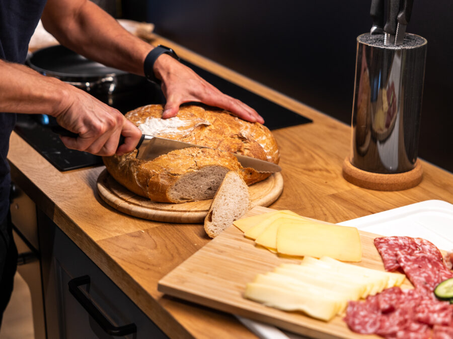 Eine Person schneidet in einer Küche einen runden Laib Brot auf einem hölzernen Schneidebrett. Daneben liegt ein Teller mit verschiedenen Käsesorten, Salami und Gurkenscheiben, und auf der Arbeitsplatte liegt ein Messerblock. Küche in einer Ferienwohnung für große Familien im Marktquartier 5 in Neusalza-Spremberg.