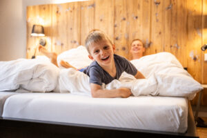 Ein kleiner Junge lächelt und liegt verspielt auf einem Bett mit weißen Laken, während sich im Hintergrund ein Erwachsener entspannt. Das Zimmer hat ein gemütliches Holzkopfteil und eine sanfte, warme Atmosphäre. Elternschlafzimmer in einer Ferienwohnung für große Familien im Marktquartier 5 in Neusalza-Spremberg.