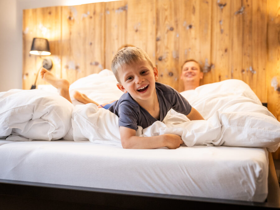Ein kleiner Junge lächelt und liegt verspielt auf einem Bett mit weißen Laken, während sich im Hintergrund ein Erwachsener entspannt. Das Zimmer hat ein gemütliches Holzkopfteil und eine sanfte, warme Atmosphäre. Elternschlafzimmer in einer Ferienwohnung für große Familien im Marktquartier 5 in Neusalza-Spremberg.