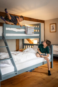 Zwei Jungen in einem Schlafzimmer mit grauen Etagenbetten; ein Junge klettert auf das obere Bett, während der andere auf dem unteren sitzt, ein Buch in der Hand hält und zu ihm aufschaut. Das Zimmer hat Holzboden und eine gemütliche Beleuchtung. Kinderzimmer in einer Ferienwohnung im Marktquartier 5 in Sachsen.