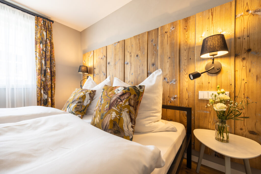 Ein gemütliches Schlafzimmer mit einem Doppelbett, weißer Bettwäsche, dekorativen Kissen mit Blumenmuster, einem hölzernen Kopfteil, Wandlampen, einem kleinen weißen Beistelltisch und einer Blumenvase neben einem Fenster mit Vorhang.