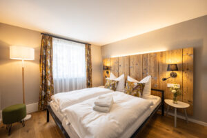 Ein gemütliches Schlafzimmer mit einem Doppelbett, weißer Bettwäsche und einem hölzernen Kopfteil. Geblümte Vorhänge am Fenster, eine Stehlampe, ein kleiner runder Tisch mit Blumen und zwei Kissen mit Blumenmustern. Warmes Licht sorgt für eine einladende Atmosphäre.