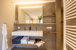 Modernes Badezimmer mit großem Spiegel, Doppelwaschbecken, Handtüchern, Haartrockner und Toilettenartikeln, ordentlich auf Holzregalen angeordnet, mit warmer Beleuchtung und Fliesen in neutralen Farben.