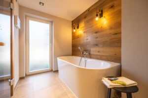 Modernes Badezimmer mit weißer freistehender Badewanne, Holzakzentwand, zwei warmen Wandleuchten, großer Milchglastür und gefalteten Handtüchern mit Toilettenartikeln auf einem kleinen Tisch. Der Raum ist sanft und warm beleuchtet.