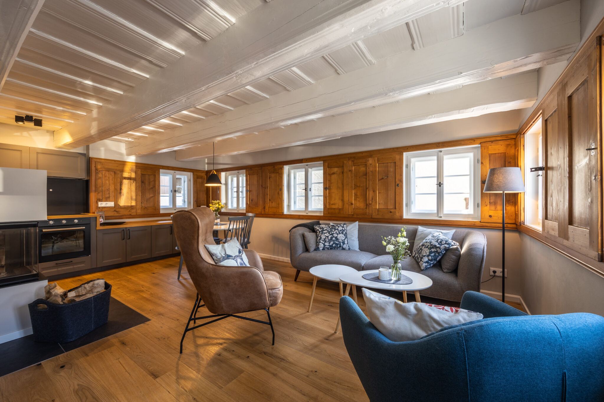 Gemütliches, modernes Wohnzimmer mit Holzboden, Sichtbalken und Holzfensterläden in einer Ferienwohnung in der Oberlausitz. Es verfügt über Sessel, Sofa, Couchtische und eine Stehlampe. Im Hintergrund befinden sich Küche und Essbereich mit Pendelleuchte. Wohnzimmer im Apartment Alwine im Ferienhaus Marktquartier 5.