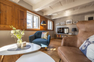 Ein gemütliches Wohnzimmer mit Holzboden, einem blauen Sessel, einem braunen Hochlehner, einem weißen runden Tisch mit Blumen, hölzernen Fensterläden und einem Fernseher auf einem Ständer mit Büchern darunter. Sonnenlicht strömt durch die Fenster.