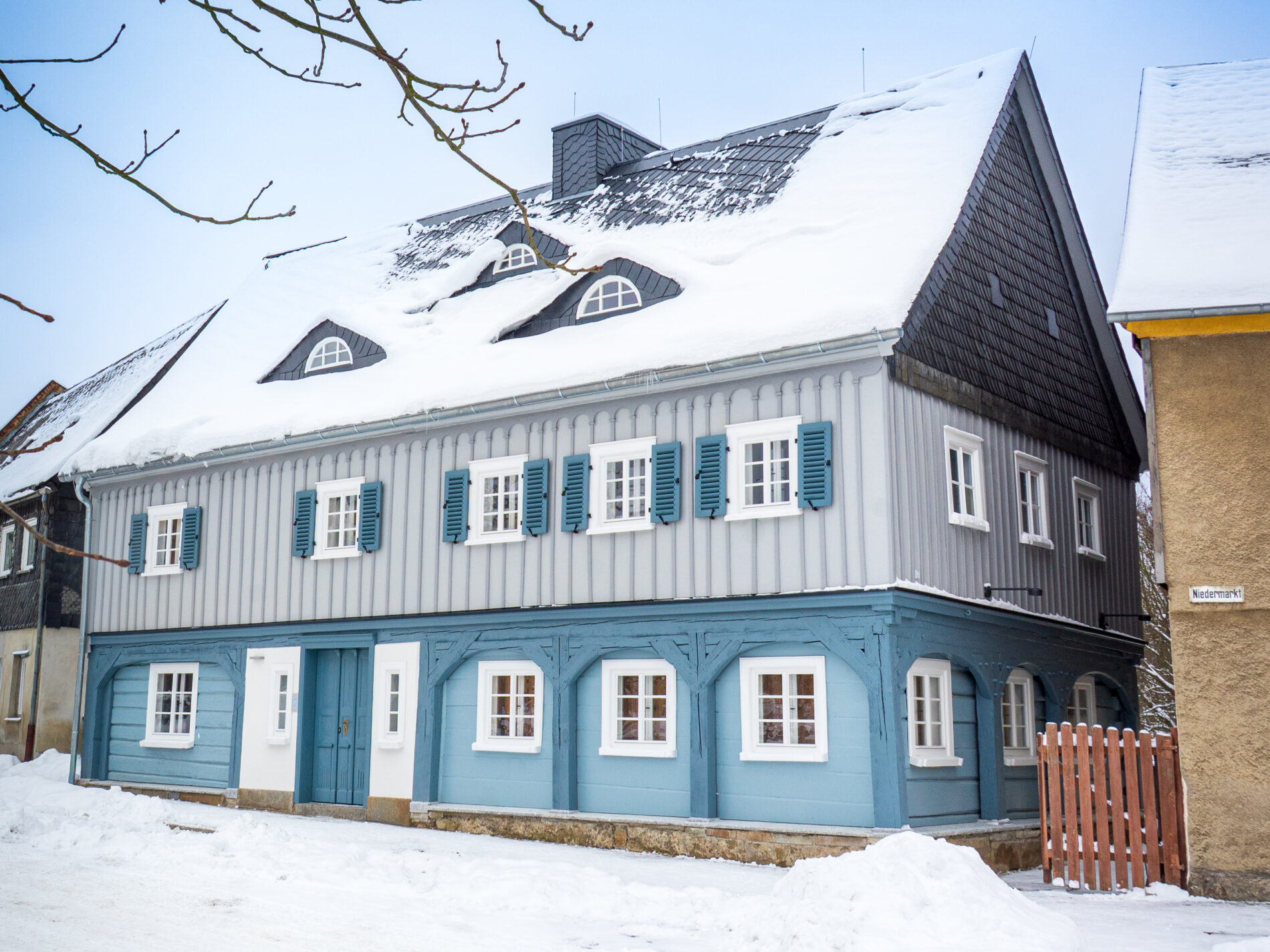 Ferienhaus Marktquartier 5 - ein traditionelles, zweistöckiges, blau-graues Umgebindehaus in Neusalza-Spremberg, dessen Dach und Boden mit Schnee bedeckt sind. Das Haus hat weiß gerahmte Fenster mit blauen Fensterläden und dekorative Holzarbeiten an der Fassade. Vor dem Haus sind kahle Baumzweige zu sehen.