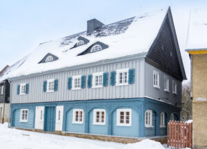 Ferienhaus Marktquartier 5 - ein zweistöckiges blau-graues Umgebindehaus in Neusalza-Spremberg, dessen Dach und Fensterläden mit Schnee bedeckt sind, umgeben von Schnee auf dem Boden, unter einem bewölkten Winterhimmel.