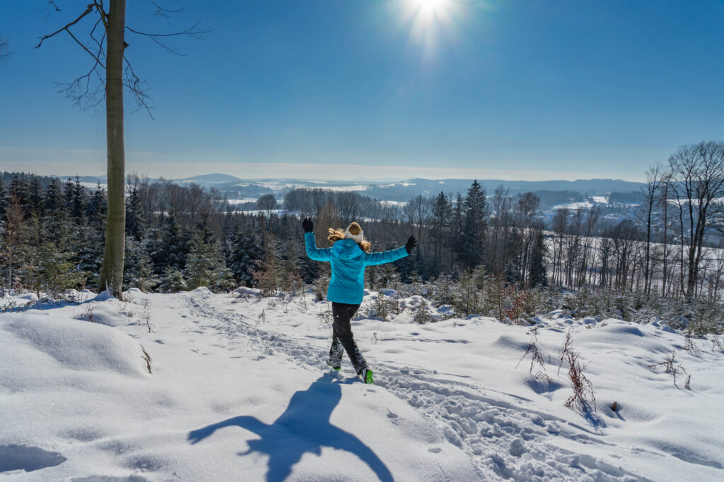 Eine Person in leuchtend blauer Jacke läuft durch tiefen Schnee in einer sonnigen Winterlandschaft, umgeben von Bäumen und Hügeln unter einem klaren Himmel - perfekt für Inspiration und Tipps für Winterurlaub in der Oberlausitz.