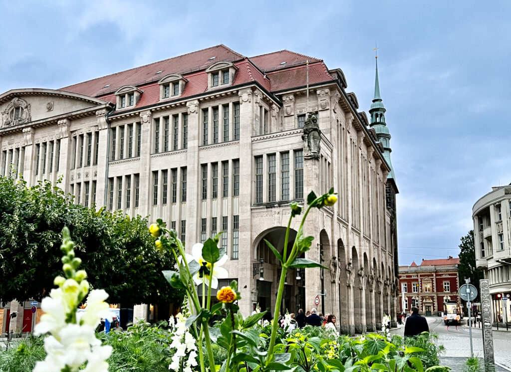 Das Kaufhaus in Görlitz. Ein prächtiges historisches Gebäude mit hohen Bogenfenstern und einem roten Ziegeldach steht hinter einem Blumengarten. Bekannt als einer der Filmdrehorte in der Oberlausitz, erhebt sich auf der rechten Seite ein grüner Kirchturm unter einem bewölkten Himmel.
